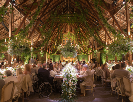A vibrant, lush wedding reception in the Old Barn of The Santa Lucia Preserve. Tables full of wedding guests fill the space, with large leafy chandeliers and ivy hanging from the 3-story tall ceiling.
